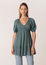 [Color: Lagoon] A front facing image of a blonde model wearing a bohemian spring tunic top in a teal crinkled fabric. With short puff sleeves, ruffle trim, a v neckline, a smocked bodice, an empire waist, and a flowy tiered body. 