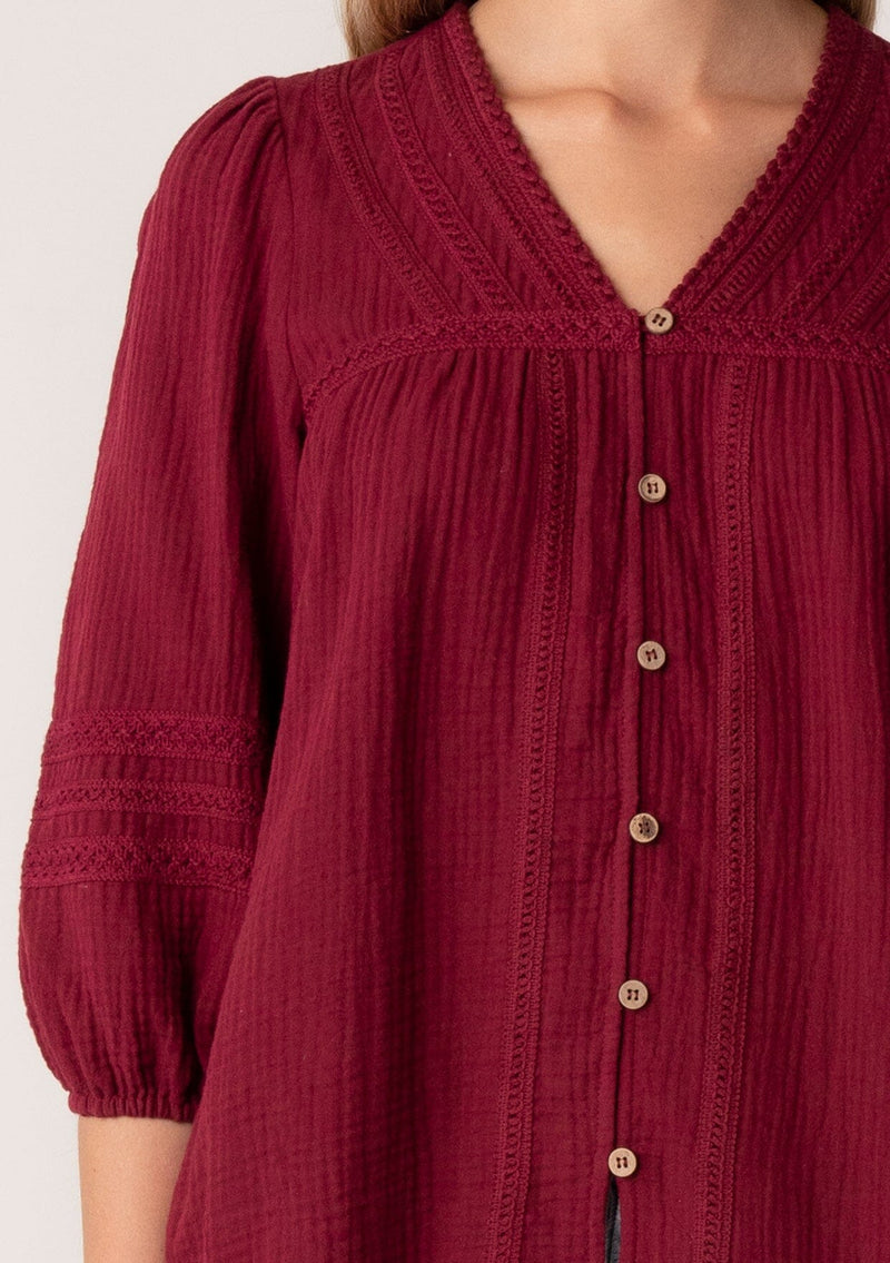 [Color: Merlot] A close up front facing image of a blonde model wearing a classic bohemian blouse in a burgundy red soft cotton gauze. With three quarter length puff sleeves, a v neckline, a button front, and embroidered details throughout. 