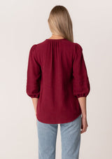 [Color: Merlot] A back facing image of a blonde model wearing a classic bohemian blouse in a burgundy red soft cotton gauze. With three quarter length puff sleeves, a v neckline, a button front, and embroidered details throughout. 