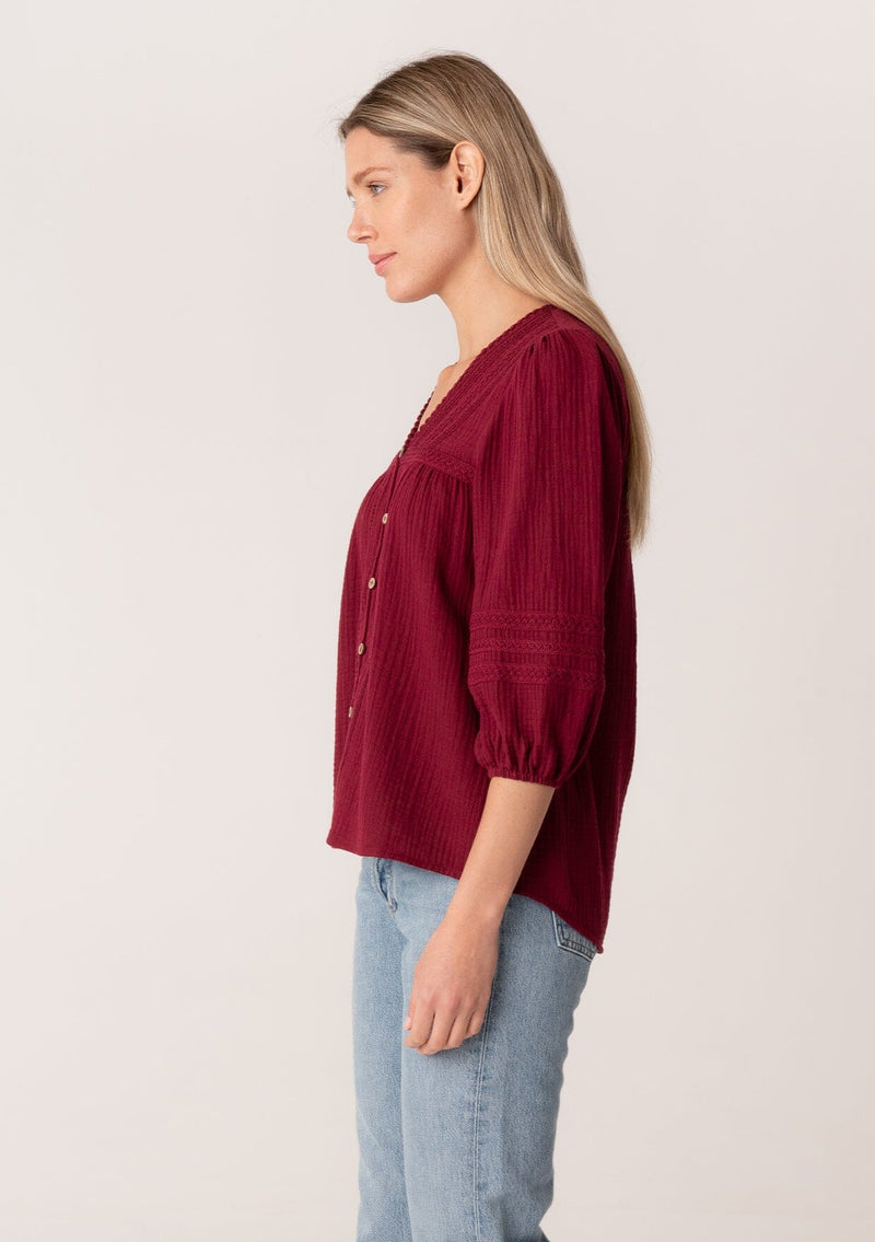 [Color: Merlot] A side facing image of a blonde model wearing a classic bohemian blouse in a burgundy red soft cotton gauze. With three quarter length puff sleeves, a v neckline, a button front, and embroidered details throughout. 