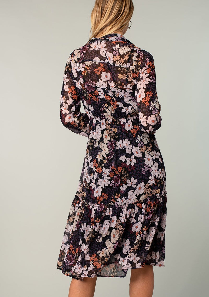 [Color: Black/Dusty Rose] A back facing image of a blonde model wearing a sheer chiffon mid length dress in a black and lavender purple floral print. With sheer long sleeves, a button front, a collared neckline, and a drawstring waist that can be adjusted to fit loose or defined. 