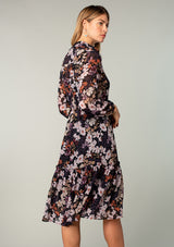[Color: Black/Dusty Rose] A back facing image of a blonde model wearing a sheer chiffon mid length dress in a black and lavender purple floral print. With sheer long sleeves, a button front, a collared neckline, and a drawstring waist that can be adjusted to fit loose or defined. 