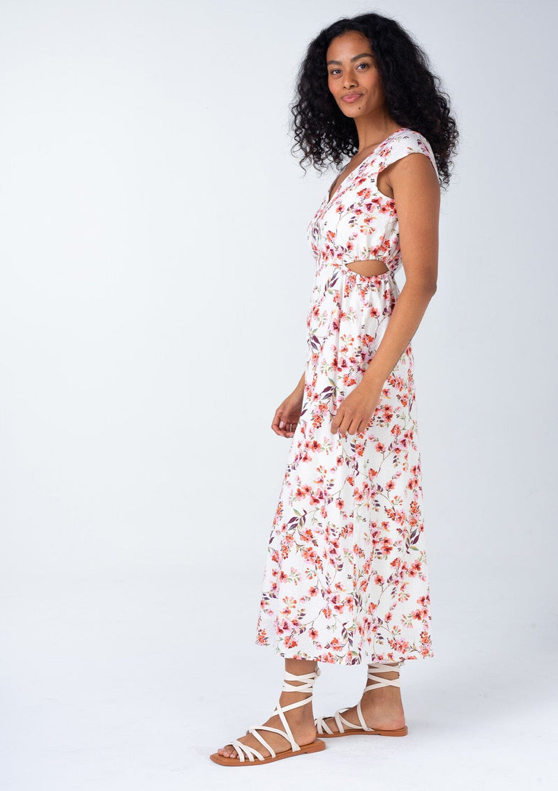 [Color: Natural/Rose] A side facing image of a brunette model wearing a cotton blend mid length spring dress in a natural and rose pink floral print. With short cap sleeves, a self covered button front, a v neckline, and sexy side waist cutouts.