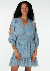 [Color: Dusty Blue/Natural] A half body front facing image of a brunette model wearing a sheer chiffon bohemian mini dress in an abstract squiggle print. With long billowy split sleeves, an open back with tassel tie closure, a smocked elastic waist, and a ruffle trimmed tiered skirt.
