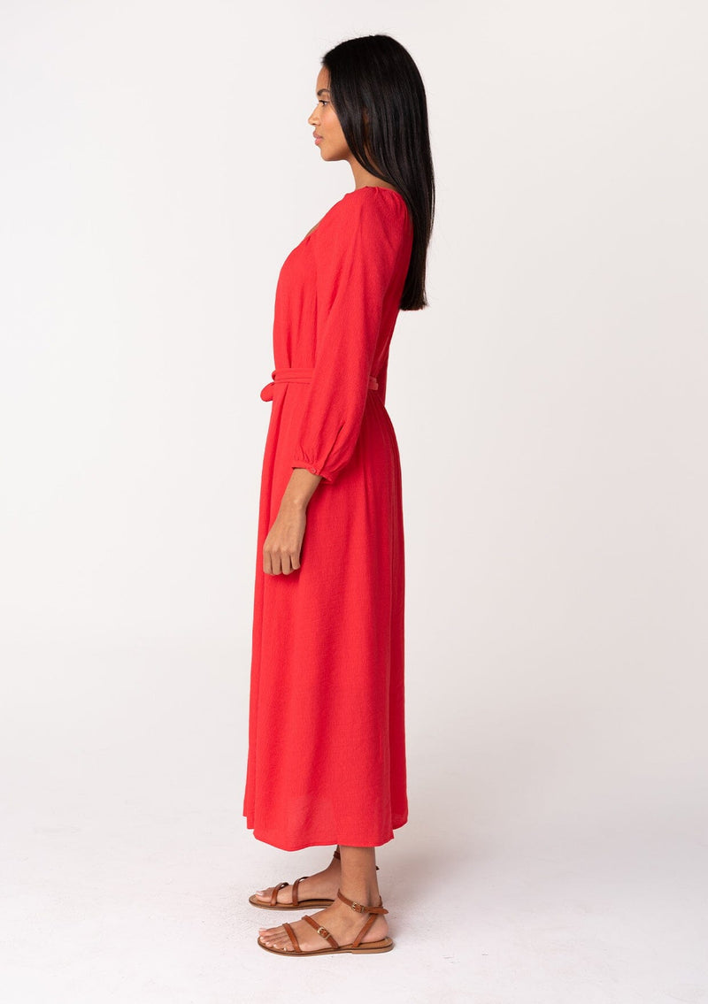 [Color: Red] A side facing image of a brunette model wearing a classic bright red bohemian maxi dress. With long sleeves, a round neckline, a button front, side pockets, and a self tie waist belt.
