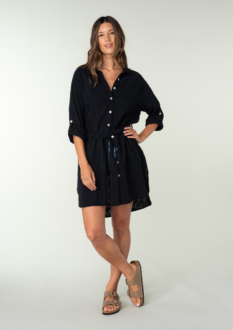 [Color: Black] A full body front facing image of a brunette model wearing a bohemian black cotton tunic shirt in a textured jacquard. With long rolled sleeves and a button tab closure, a collared neckline, a button front, a self tie waist belt, side vents, a round hemline, and a back yoke detail.