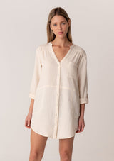 [Color: Cloud] A front facing image of a blonde model wearing a textured airy gauze light cream button front shirt. With a long tunic length, long rolled sleeves with a button tab, a single front pocket, and a banded collar with a v neckline.
