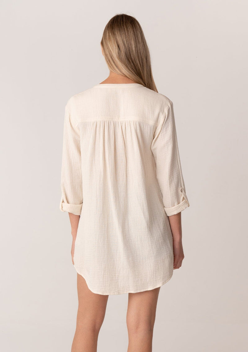 [Color: Cloud] A back facing image of a blonde model wearing a textured airy gauze light cream button front shirt. With a long tunic length, long rolled sleeves with a button tab, a single front pocket, and a banded collar with a v neckline.