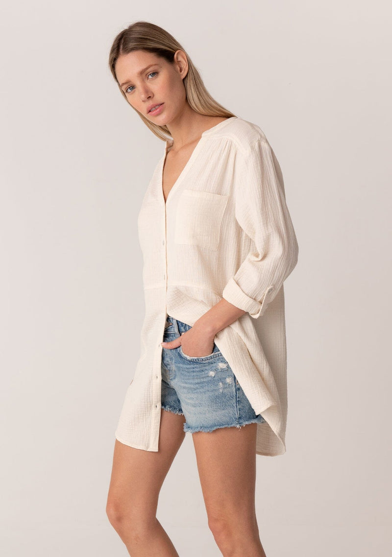[Color: Cloud] A side facing image of a blonde model wearing a textured airy gauze light cream button front shirt. With a long tunic length, long rolled sleeves with a button tab, a single front pocket, and a banded collar with a v neckline.