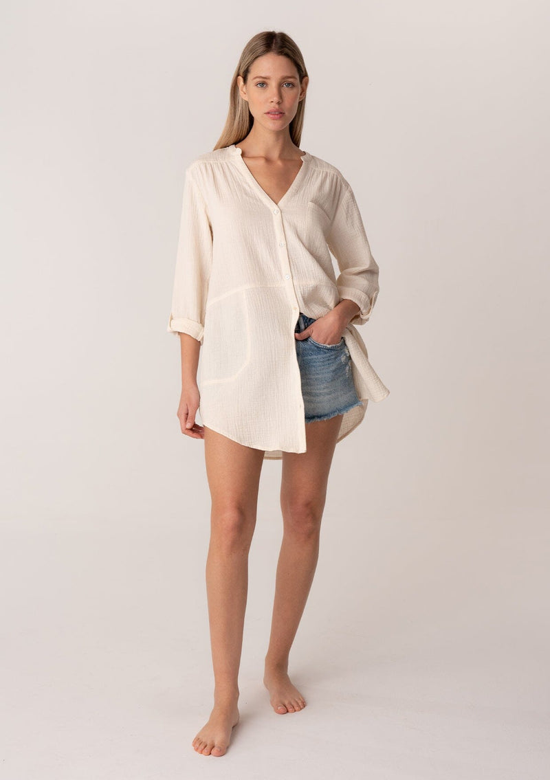 [Color: Cloud] A full body front facing image of a blonde model wearing a textured airy gauze light cream button front shirt. With a long tunic length, long rolled sleeves with a button tab, a single front pocket, and a banded collar with a v neckline.