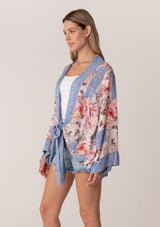 [Color: Natural/Periwinkle] A side facing image of a blonde model wearing a bohemian kimono top in a blue and pink floral print. With long flared sleeves, a relaxed flowy fit, and a tie front waist. 