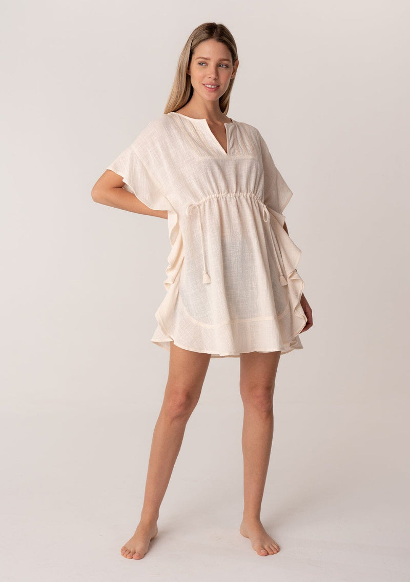 [Color: Natural] A full body front facing image of a blonde model wearing a bohemian caftan top in an off white cotton. A beach cover up style with short sleeves, a ruffled hemline, a v neckline, embroidered details throughout, and a drawstring waist detail in the front and back with tassel ties. 