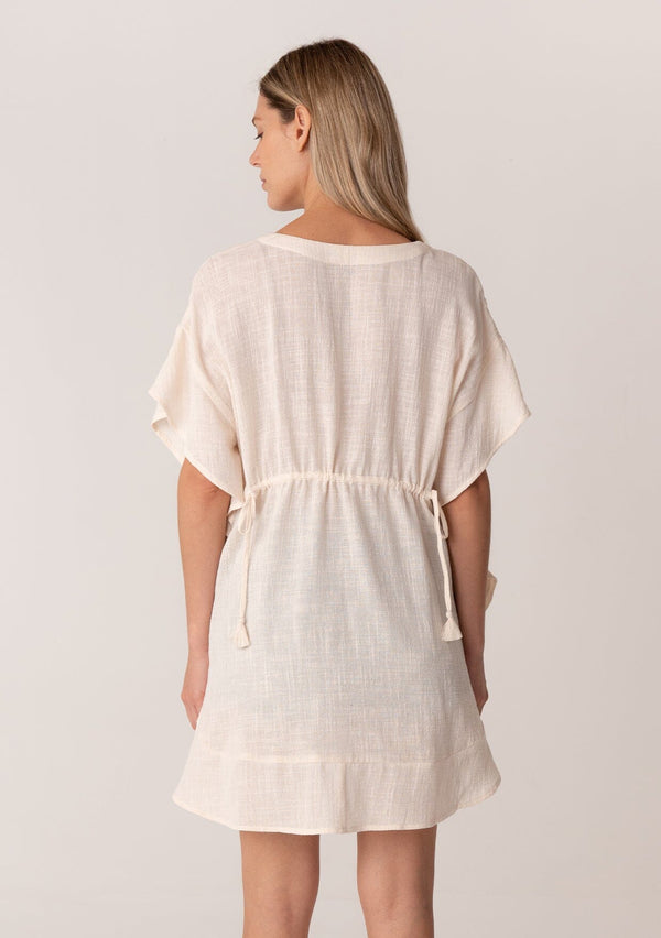 [Color: Natural] A back facing image of a blonde model wearing a bohemian caftan top in an off white cotton. A beach cover up style with short sleeves, a ruffled hemline, a v neckline, embroidered details throughout, and a drawstring waist detail in the front and back with tassel ties. 
