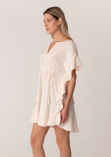 [Color: Natural] A side facing image of a blonde model wearing a bohemian caftan top in an off white cotton. A beach cover up style with short sleeves, a ruffled hemline, a v neckline, embroidered details throughout, and a drawstring waist detail in the front and back with tassel ties. 