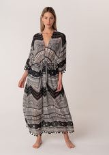 [Color: Black/Natural] A full body front facing image of a blonde model wearing a black printed bohemian resort kimono. A maxi length cover up with tassel fringe hem, half length sleeves, and a tie waist belt. 