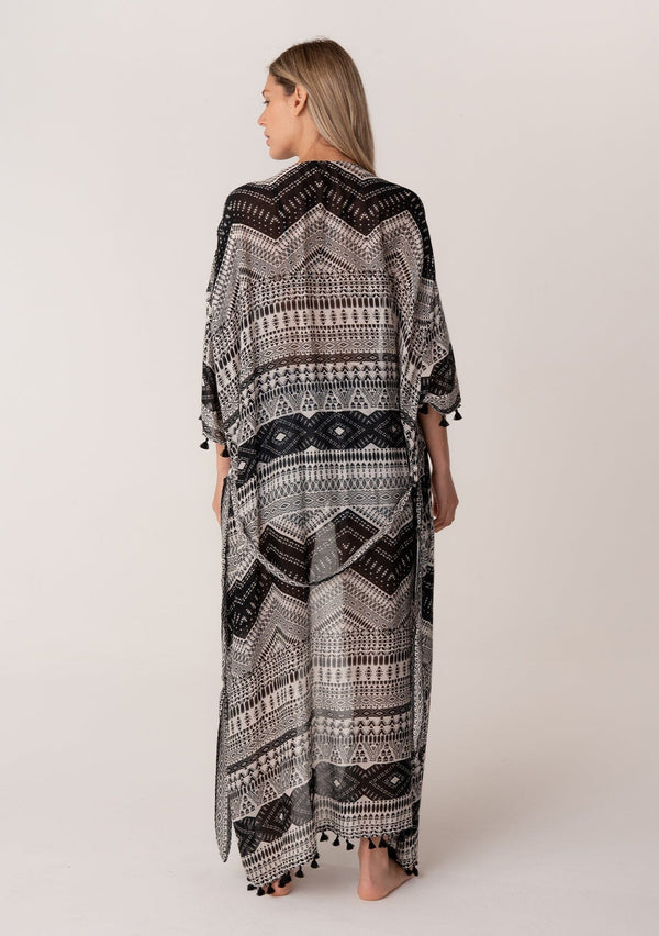 [Color: Black/Natural] A back facing image of a blonde model wearing a black printed bohemian resort kimono. A maxi length cover up with tassel fringe hem, half length sleeves, and a tie waist belt. 