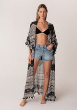 [Color: Black/Natural] A front facing image of a blonde model wearing a black printed bohemian resort kimono. A maxi length cover up with tassel fringe hem, half length sleeves, and a tie waist belt. 