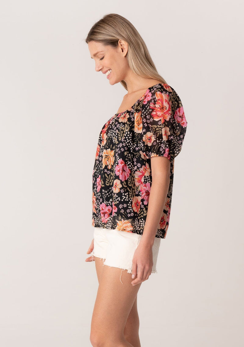 [Color: Black/Fuchsia] A side facing image of a blonde model wearing a bohemian spring top in a black and pink floral print. With short puff sleeves, a square neckline, and a relaxed fit. 
