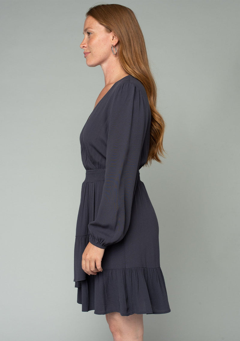 [Color: Charcoal] A side facing image of a red headed model wearing a charcoal grey bohemian mini dress designed in a lightweight crepe. With long sleeves, a surplice v neckline, and a ruffled faux wrap skirt. 