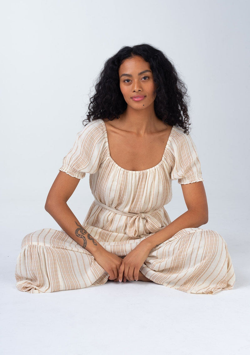[Color: Natural/Tan] A sitting front facing image of a brunette model wearing a flowy bohemian resort maxi dress in a neutral natural and tan stripe. With short puff sleeves, a rounded square neckline, a tiered skirt, a self tie waist belt, and a tassel tie drawstring detail at the back.