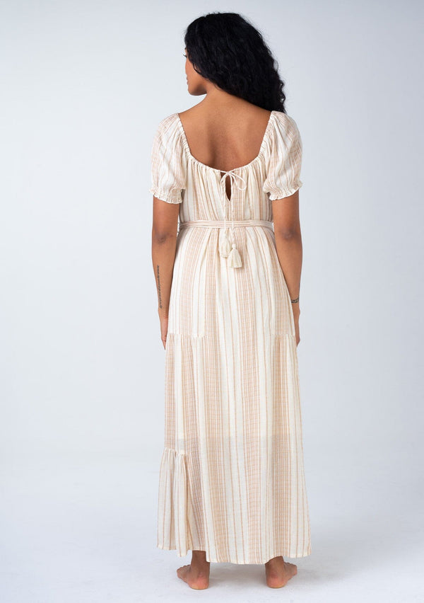 [Color: Natural/Tan] A back facing image of a brunette model wearing a flowy bohemian resort maxi dress in a neutral natural and tan stripe. With short puff sleeves, a rounded square neckline, a tiered skirt, a self tie waist belt, and a tassel tie drawstring detail at the back.