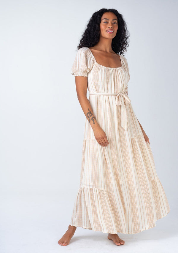 [Color: Natural/Tan] A front facing image of a brunette model wearing a flowy bohemian resort maxi dress in a neutral natural and tan stripe. With short puff sleeves, a rounded square neckline, a tiered skirt, a self tie waist belt, and a tassel tie drawstring detail at the back.