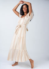 [Color: Natural/Tan] A full body side facing image of a brunette model wearing a flowy bohemian resort maxi dress in a neutral natural and tan stripe. With short puff sleeves, a rounded square neckline, a tiered skirt, a self tie waist belt, and a tassel tie drawstring detail at the back.