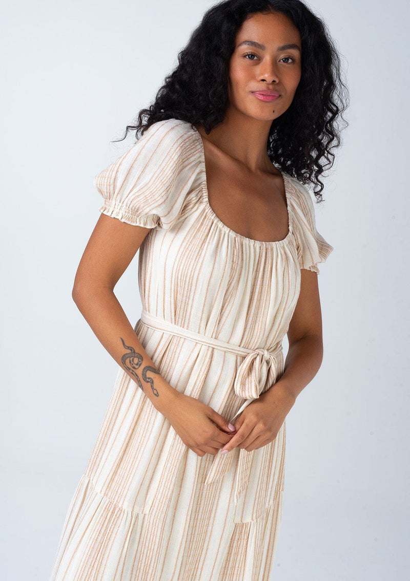 [Color: Natural/Tan] A close up front facing image of a brunette model wearing a flowy bohemian resort maxi dress in a neutral natural and tan stripe. With short puff sleeves, a rounded square neckline, a tiered skirt, a self tie waist belt, and a tassel tie drawstring detail at the back.