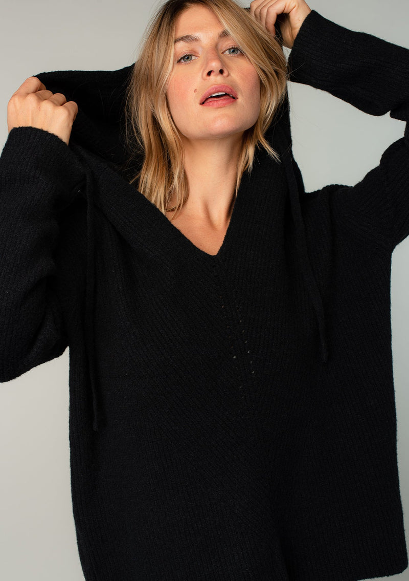 [Color: Black] A model wearing a black cozy knit pullover sweater. Featuring a drawstring hoodie, long sleeves, a v neckline, and open knit details. A classic Baja sweater silhouette.