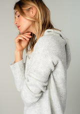 [Color: Light Heather Grey] A model wearing a light heather grey cozy knit pullover sweater. Featuring a drawstring hoodie, long sleeves, a v neckline, and open knit details. A classic Baja sweater silhouette.