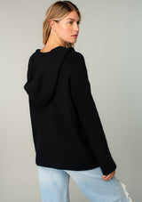 [Color: Black] A model wearing a black cozy knit pullover sweater. Featuring a drawstring hoodie, long sleeves, a v neckline, and open knit details. A classic Baja sweater silhouette.