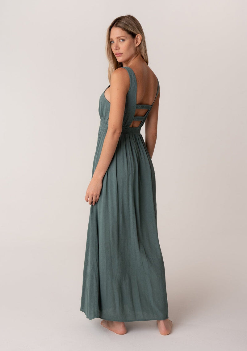 [Color: Lagoon] A full body back facing image of a blonde model wearing a bohemian sleeveless spring maxi dress in dusty teal. With a self covered button front, a deep v neckline, a front slit, a smocked elastic empire waist, and an open back with elastic strap detail.
