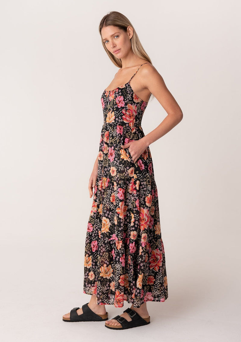 [Color: Black/Fuchsia] A side facing image of a blonde model wearing a pretty spring maxi dress in a black and pink floral print. With spaghetti straps, a scooped neckline, a flowy tiered skirt, a button front, and a half smocked elastic bodice at the back. 