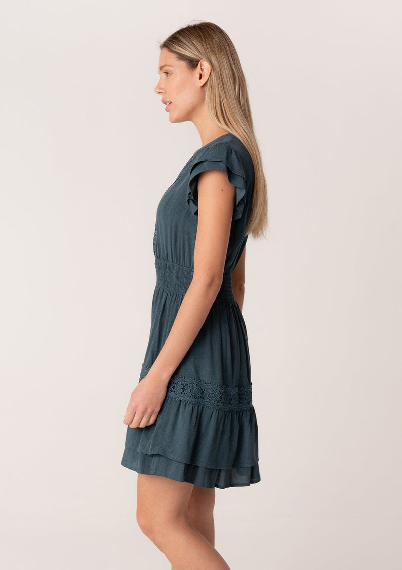 [Color: Teal] A side facing image of a blonde model wearing a teal blue bohemian mini dress with short double flutter sleeves, a smocked elastic waist, a double layered skirt, a split v neckline, and lace trim detail throughout.