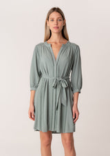 [Color: Sea Green] A front facing image of a blonde model wearing a dusty teal bohemian spring mini dress. With three quarter length long sleeves, lattice lace trim details, a concealed button front, and a self tie waist belt.
