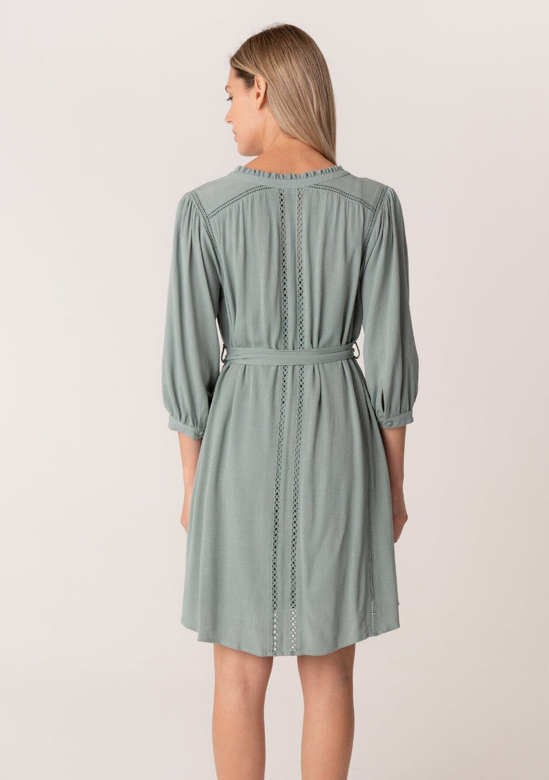 [Color: Sea Green] A back facing image of a blonde model wearing a dusty teal bohemian spring mini dress. With three quarter length long sleeves, lattice lace trim details, a concealed button front, and a self tie waist belt.