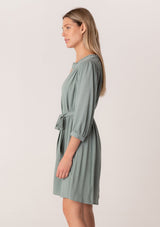 [Color: Sea Green] A side facing image of a blonde model wearing a dusty teal bohemian spring mini dress. With three quarter length long sleeves, lattice lace trim details, a concealed button front, and a self tie waist belt.