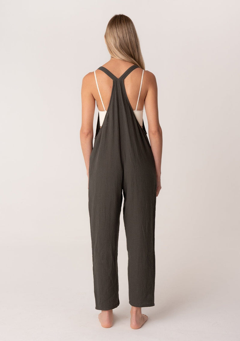 [Color: Military] A back facing image of a blonde model wearing a soft cotton gauze bohemian lounge jumpsuit in military green. With the silhouette of overalls, featuring tank top straps that attach with a button closure, two side pockets, a wide leg, and a racer back. 