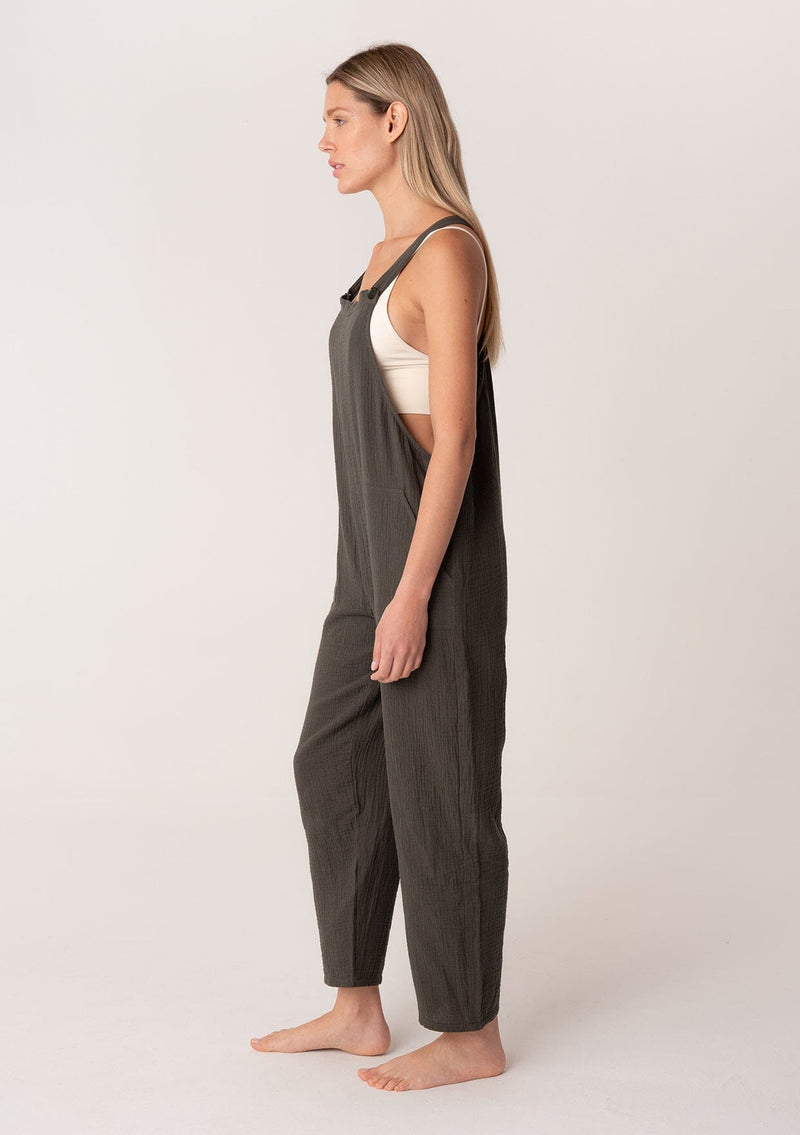 [Color: Military] A side facing image of a blonde model wearing a soft cotton gauze bohemian lounge jumpsuit in military green. With the silhouette of overalls, featuring tank top straps that attach with a button closure, two side pockets, a wide leg, and a racer back. 