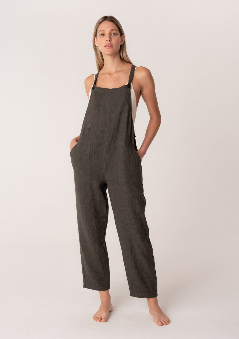 [Color: Military] A front facing image of a blonde model wearing a soft cotton gauze bohemian lounge jumpsuit in military green. With the silhouette of overalls, featuring tank top straps that attach with a button closure, two side pockets, a wide leg, and a racer back. 