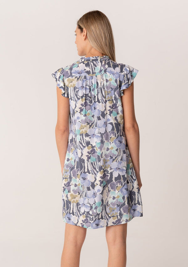 [Color: Dusty Lilac/Dusty Teal] A back facing image of a blonde model wearing a timeless spring mini dress in a lilac purple and teal floral print. With short flutter sleeves, a button front, side pockets, and a ruffled neckline.