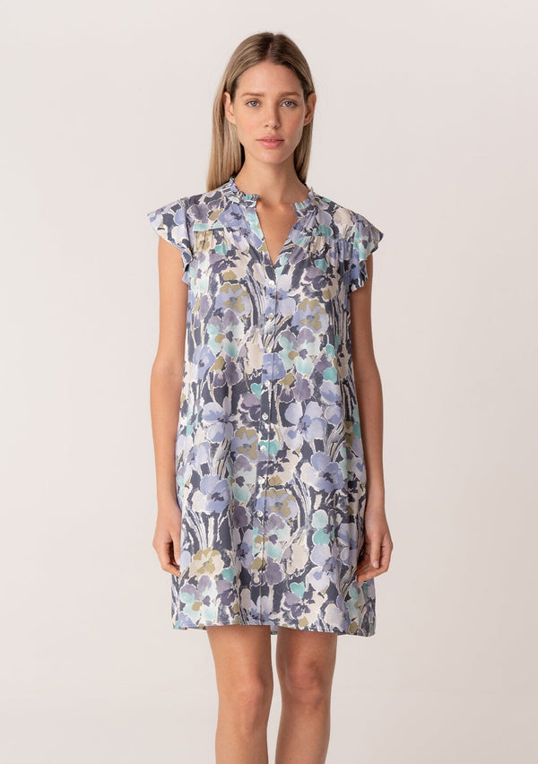 [Color: Dusty Lilac/Dusty Teal] A front facing image of a blonde model wearing a timeless spring mini dress in a lilac purple and teal floral print. With short flutter sleeves, a button front, side pockets, and a ruffled neckline.