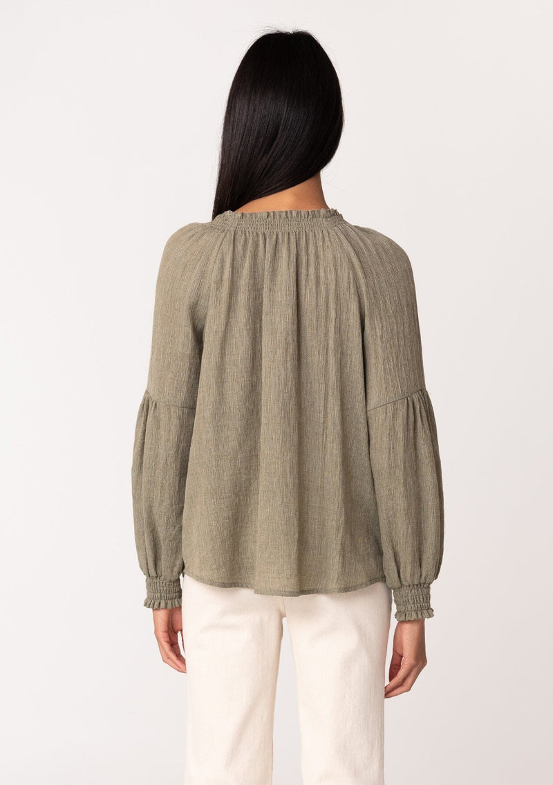 [Color: Olive] A back facing image of a brunette model wearing a classic lightweight sheer olive green peasant top. With a ruffled neckline, tassel ties, long voluminous bishop sleeves, and a relaxed flowy fit.