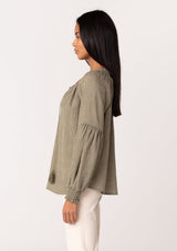 [Color: Olive] A side facing image of a brunette model wearing a classic lightweight sheer olive green peasant top. With a ruffled neckline, tassel ties, long voluminous bishop sleeves, and a relaxed flowy fit.