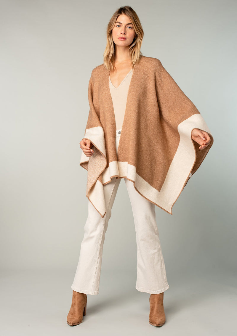 [Color: Heather Camel/Heather Ivory] A full body front facing image of a blonde model wearing a soft and warm mid length sweater cape. An open front cape cardigan with a contrast border design. Perfect fall sweater, great for layering. 