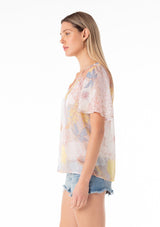 [Color: Natural/Blue] A side facing image of a blonde model wearing a pretty spring chiffon blouse designed in a pink and blue floral print. With short flutter sleeves, a split v neckline with ties, and a relaxed flowy fit. 