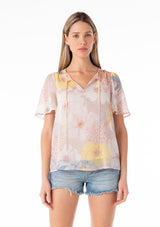 [Color: Natural/Blue] A front facing image of a blonde model wearing a pretty spring chiffon blouse designed in a pink and blue floral print. With short flutter sleeves, a split v neckline with ties, and a relaxed flowy fit. 
