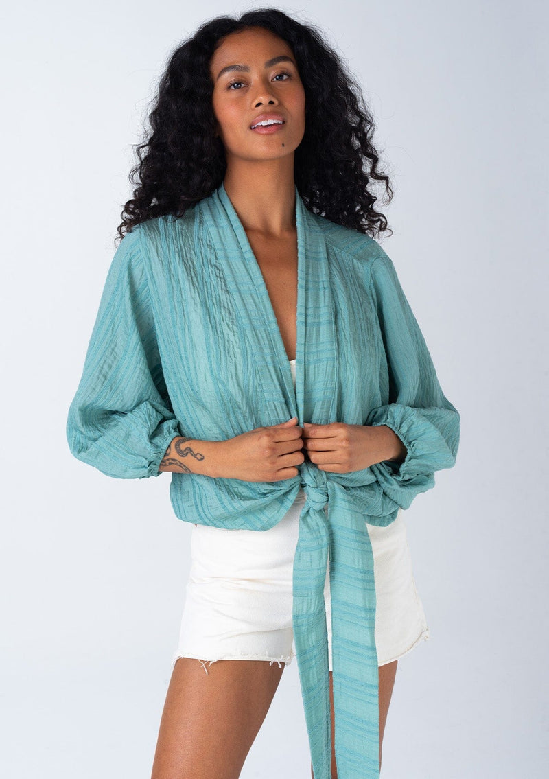 [Color: Dusty Teal] A front facing image of a brunette model wearing a best selling bohemian tie front resort top in a dusty teal shadow stripe. With voluminous long sleeves and a tie front detail that can be styled in multiple ways.