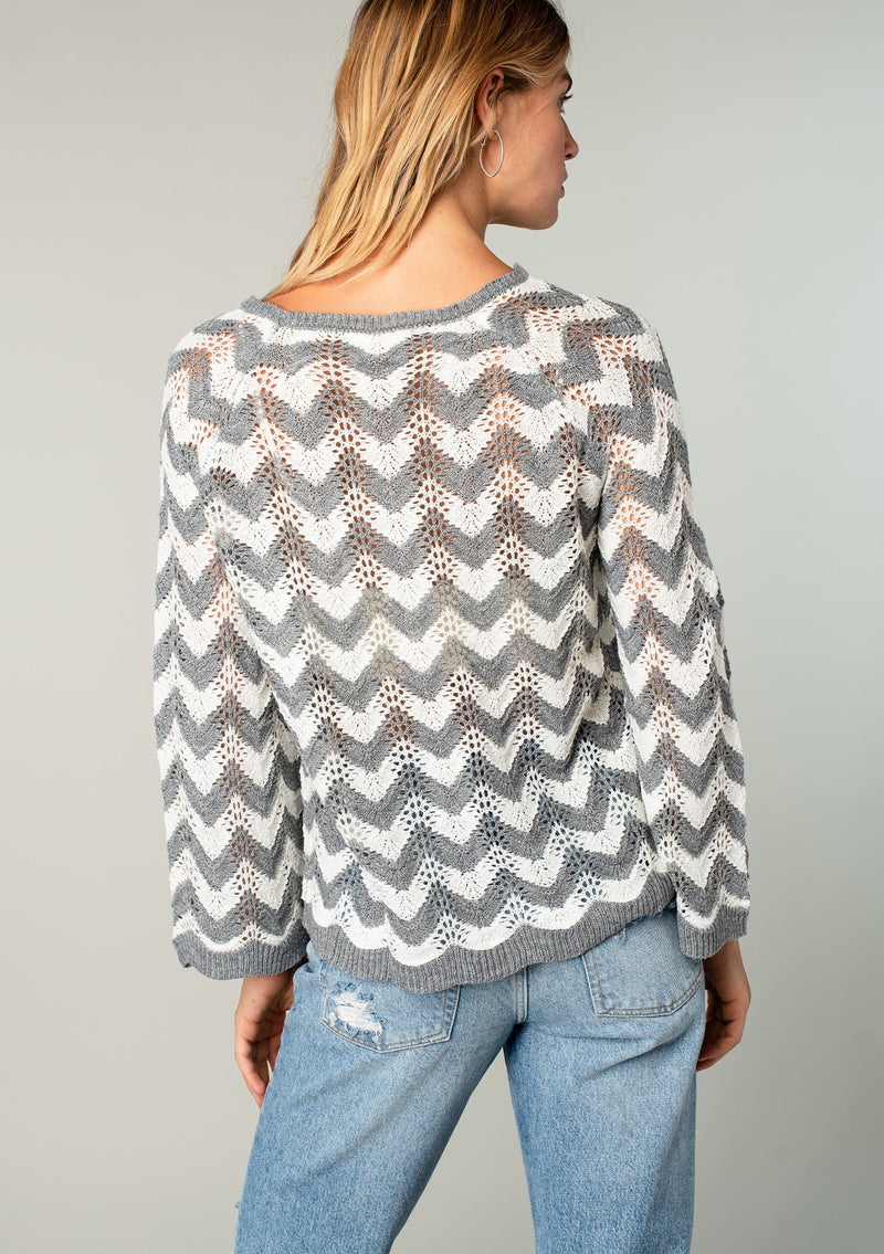 [Color: Heather Grey/Ivory] A model wearing a grey and off white crochet knit sweater with bohemian long bell sleeves and a chevron knit pattern.