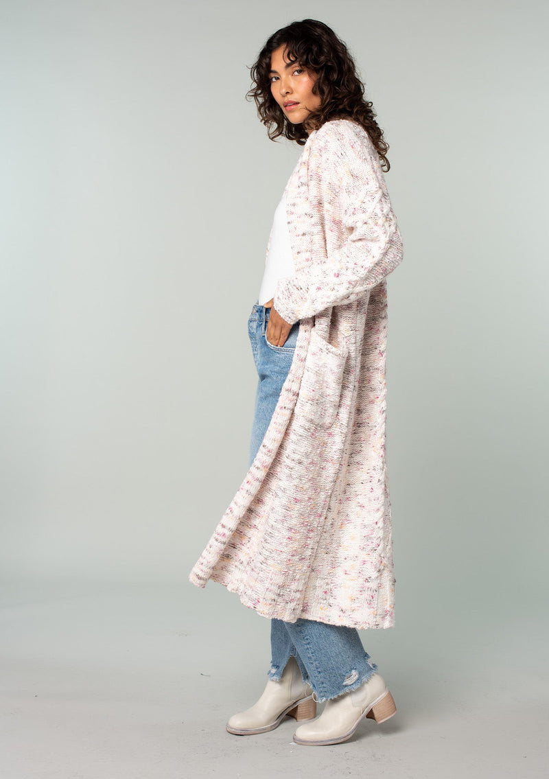 [Color: Natural/Berry] A side facing image of a brunette model wearing a natural off white and berry speckled knit long cardigan with an open front, side pockets, and cable knit details. 